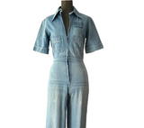 A light blue denim short-sleeve jumpsuit displayed on a mannequin in an indoor room setting with a white wall