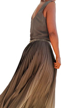 A woman wearing a sleeveless dark grey pleated maxi dress with a beaded waistband, shown from a side-back angle against a plain  wall.
