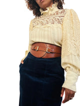 A person wearing a cream lace and ribbed blouse with a brown belt, paired with a dark navy corduroy skirt, against a plain light background.