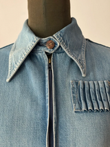 A close-up of a light blue denim jacket displayed on a dark mannequin torso. The jacket features a pointed collar with a single brown button, a central zipper, and a pleated chest pocket.