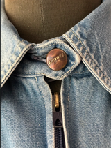 Close-up of a light blue denim jacket on a mannequin, showing a brown metal button with the brand name 'Ladder' and a dark zipper.